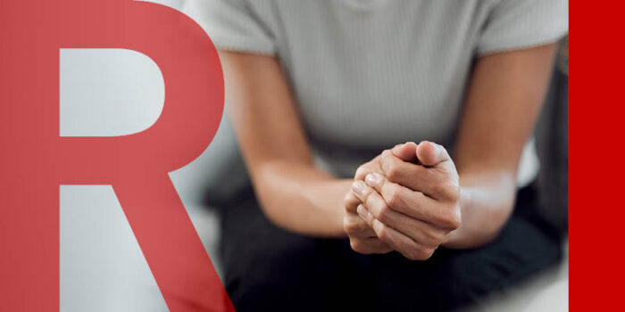 A close up of someone hands. They are sitting, leaning forward with their elbows on their knees, with their hands clasped together.