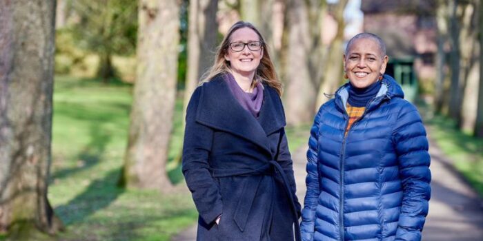 Two women standing on a path in a park with large trees behind them