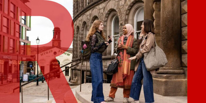Three women standing and chatting on steps outside a Victorian-style building with ornate stone architecture. The background shows the White Cloth building, pavement, and a cobbled road lined with modern shops.