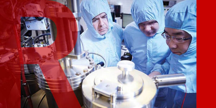 3 researchers in blue overalls looking at a metallic piece of equipment