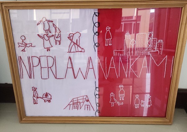 Hand embroidered artwork on half-white, half-red fabric. The left side shows people farming, dancing, and rowing. The right shows families with children and pets, and a city scape. A word in the centre reads: INIPERLAWANANKAM