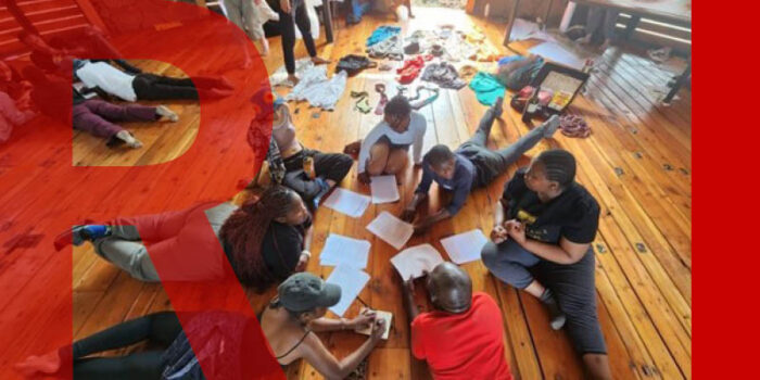 A group of young people lying on a hardwood floor in a circle, facing each other. They're working on sheets of paper with creative tools around them.