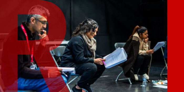 Three people sit in a dark theatre. They are leaning forward on fold-up chairs, each reading paper scripts.