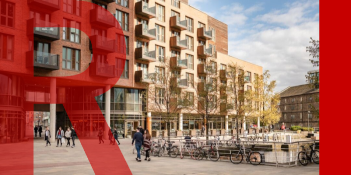 Granary Wharfe in central Leeds. Leeds Canal can be seen with a lock to the canal in the far right distance.  There are several bikes locked to the white railings which edge the canal. There are several people in groups, pairs or individuals walking in all directions through the space. Blocks of modern apartments with restaurants and cafes underneath sit in the background.